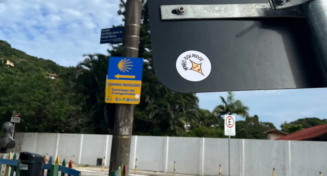 Adesivo redondo e branco colado no verso de uma placa de rua, com uma estrela amarela de quatro pontas e a frase “Honre seu poder” em volta; ao fundo, a placa azul e amarela do Caminho Brasileiro de Santiago de Compostela