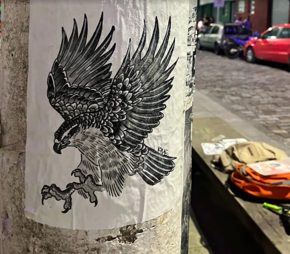 Lambe-lambe de uma águia em pleno voo, de asas abertas e garras à mostra, em preto e cinza, colado num poste de uma rua de paralelepípedos à noite