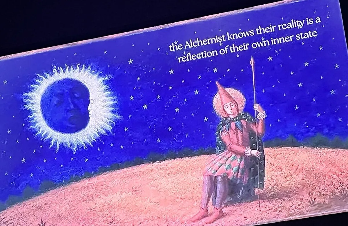 Pintura azul de noite estrelada: à esquerda, um eclipse, uma lua escura com rosto adormecido cercada por um halo de raios brancos; à direita, um arlequim de chapéu pontudo e capa verde sentado na areia, de olhos fechados, segurando uma lança. No alto, o texto em inglês “the Alchemist knows their reality is a reflection of their own inner state”