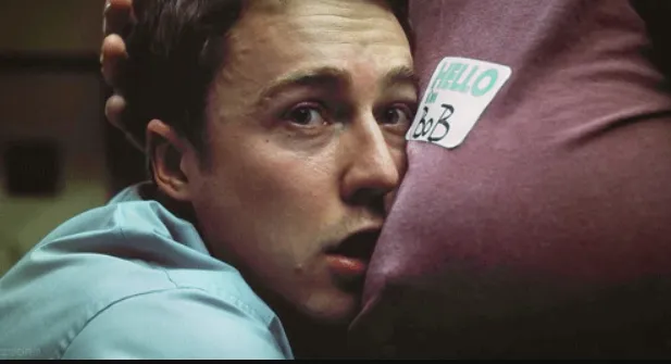 Cena do filme: um homem de camisa azul com o rosto apertado contra o peito de outro homem, que usa um adesivo escrito “HELLO my name is Bob”