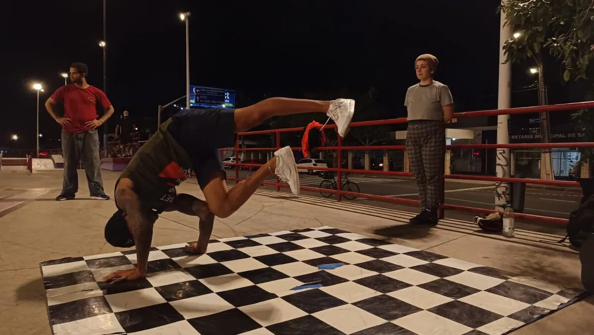 À noite, numa praça iluminada por postes, um dançarino faz um movimento de breaking apoiado nas mãos sobre um tapete xadrez preto e branco, com as pernas no ar, enquanto duas pessoas assistem em pé perto de um guarda-corpo vermelho