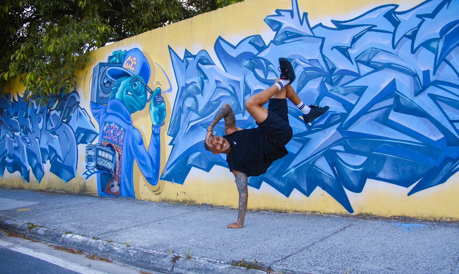 Pessoa de camiseta preta e meias brancas fazendo um freeze de breaking, apoiada numa mão só, com as pernas para o alto e sorrindo, diante de um muro amarelo grafitado de azul, ao lado do desenho de um camaleão de boné com um rádio no ombro e uma lata de spray na mão
