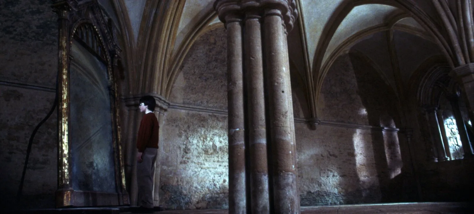 Cena do filme: Harry, pequeno, de pé diante de um espelho alto de moldura dourada, num salão de pedra vazio com colunas e arcos góticos