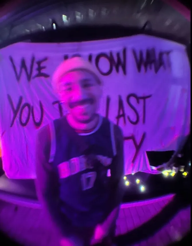 Foto embaçada sob luz roxa, com lente olho de peixe: uma pessoa de touca clara e regata de basquete sorrindo diante de um pano branco pintado com “WE KNOW WHAT YOU DID LAST…”