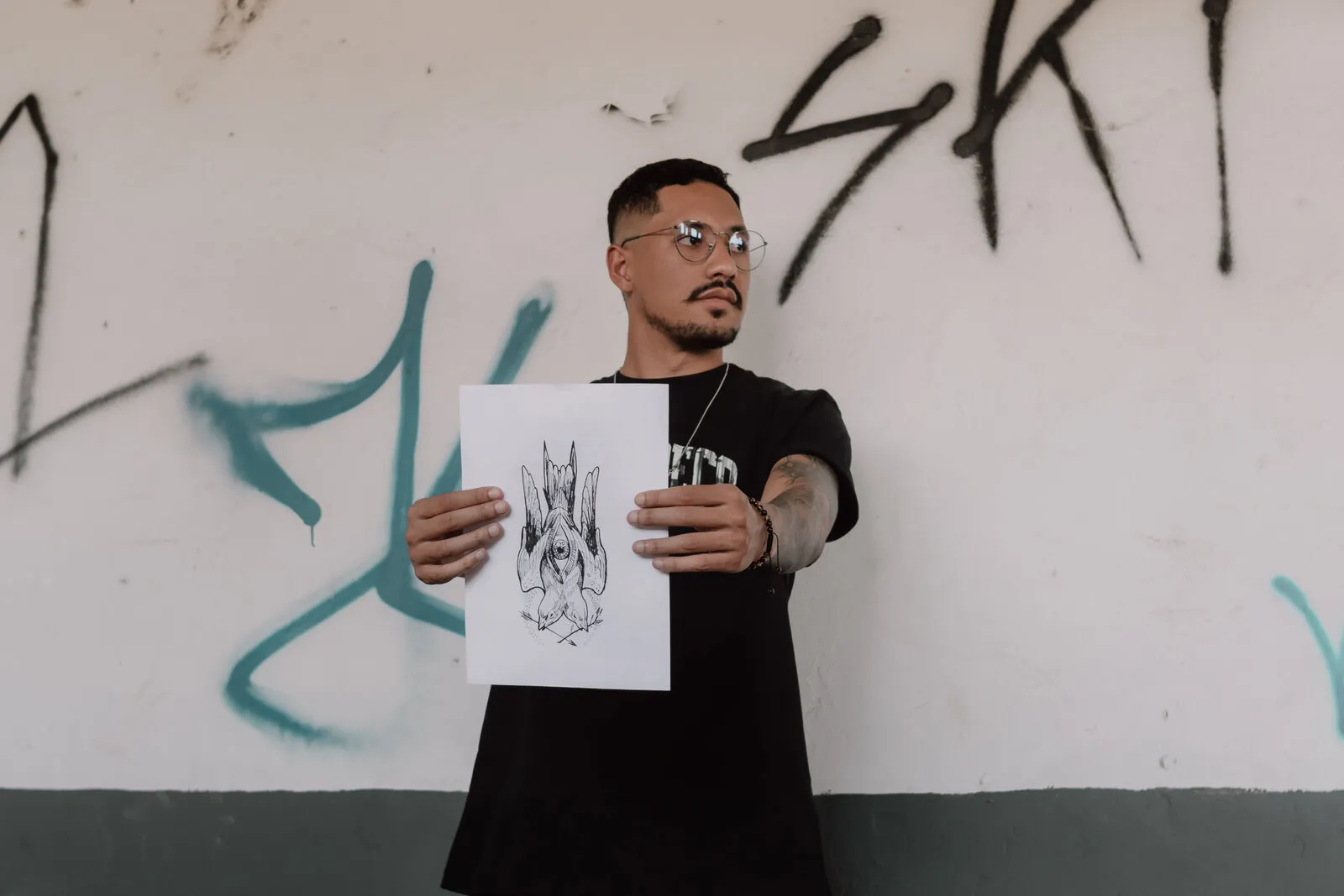 Rapaz de óculos e camiseta preta segurando um desenho em preto e branco diante de uma parede branca pichada