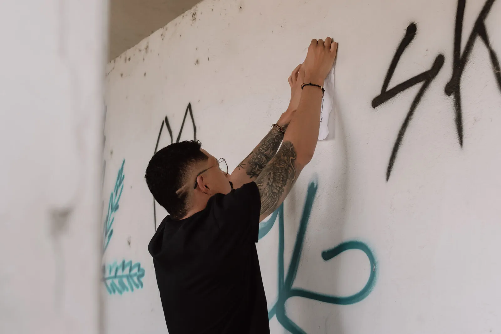 Rapaz tatuado de camiseta preta colando um papel no alto de uma parede branca pichada