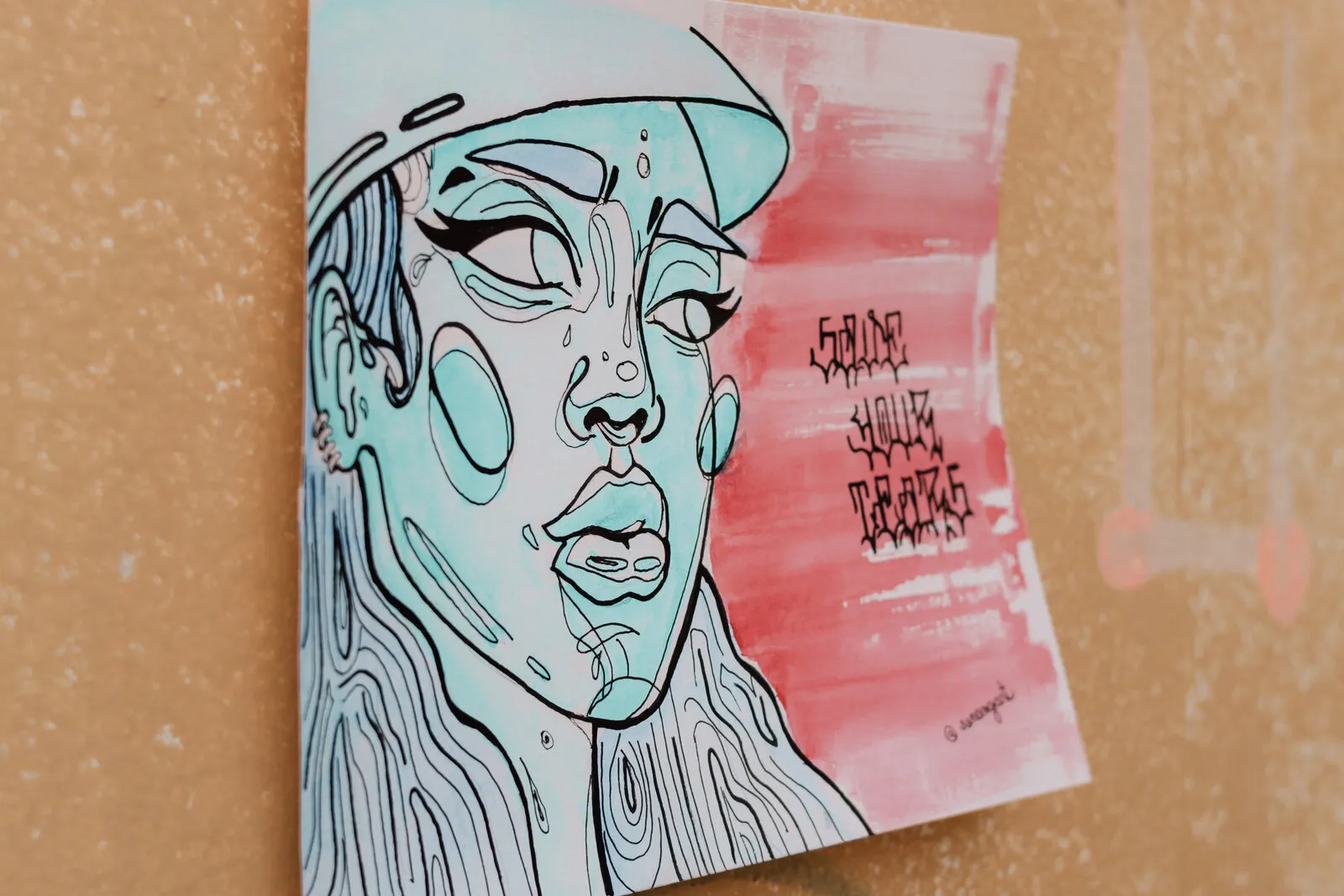 Lambe-lambe de um rosto de mulher em azul-turquesa, de boné, ao lado de uma mancha rosa com as palavras “Save your tears”, colado numa parede amarela