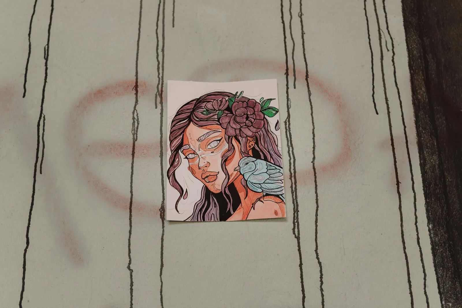 Lambe-lambe de uma mulher de cabelo roxo com flores e um passarinho no ombro, colado numa parede verde-clara com escorridos pretos e um grafite rosa apagado