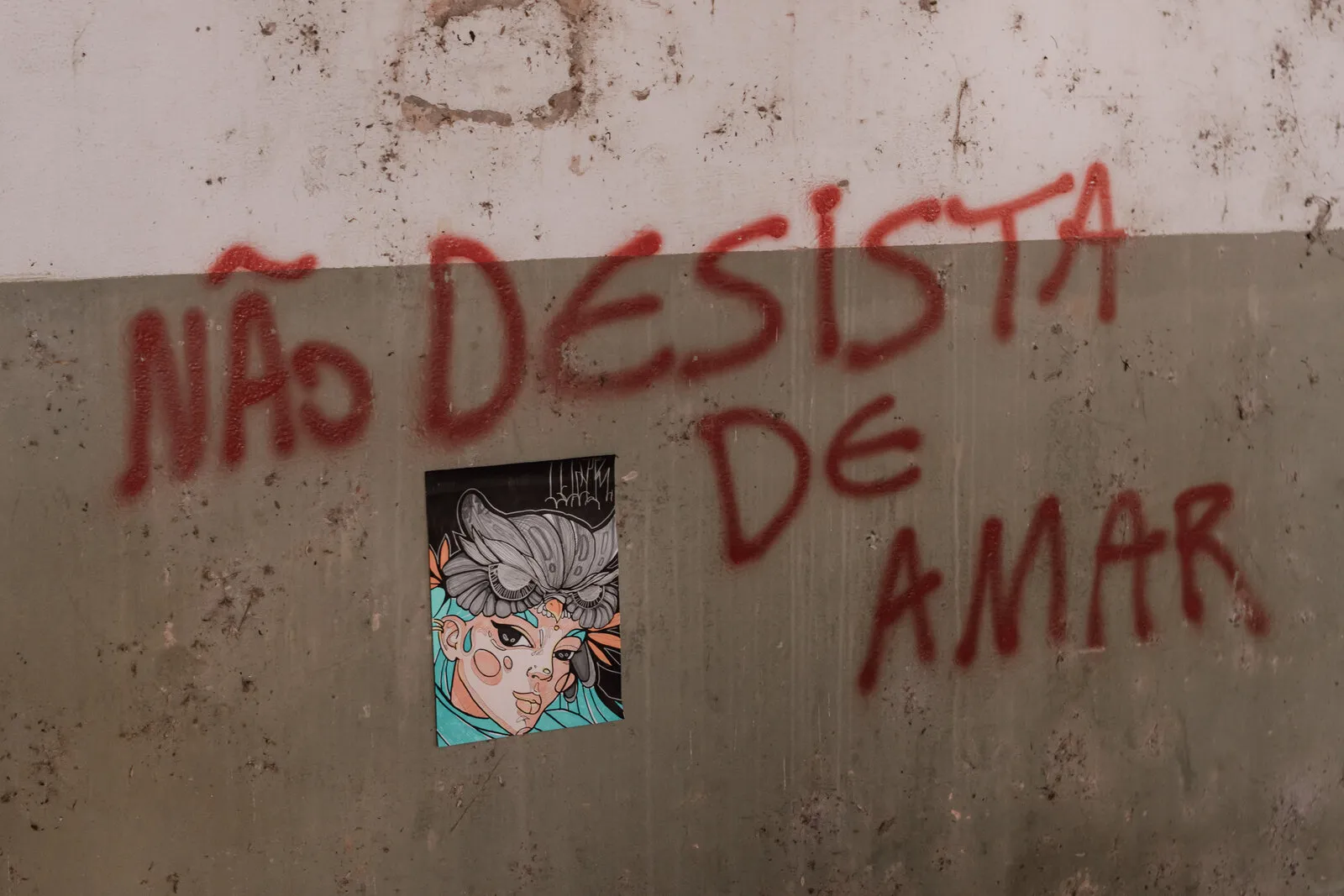 Pichação vermelha “Não desista de amar” numa parede de concreto, com um lambe-lambe de um rosto colorido colado ao lado