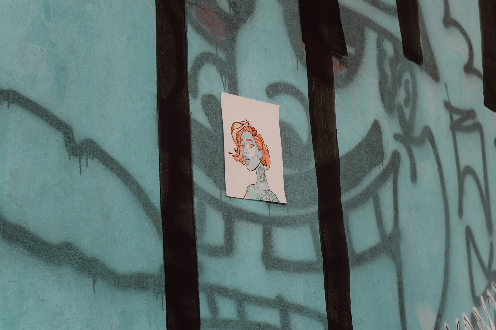 Pequeno lambe-lambe de uma mulher ruiva colado numa parede azul-turquesa com grafite preto e faixas escuras