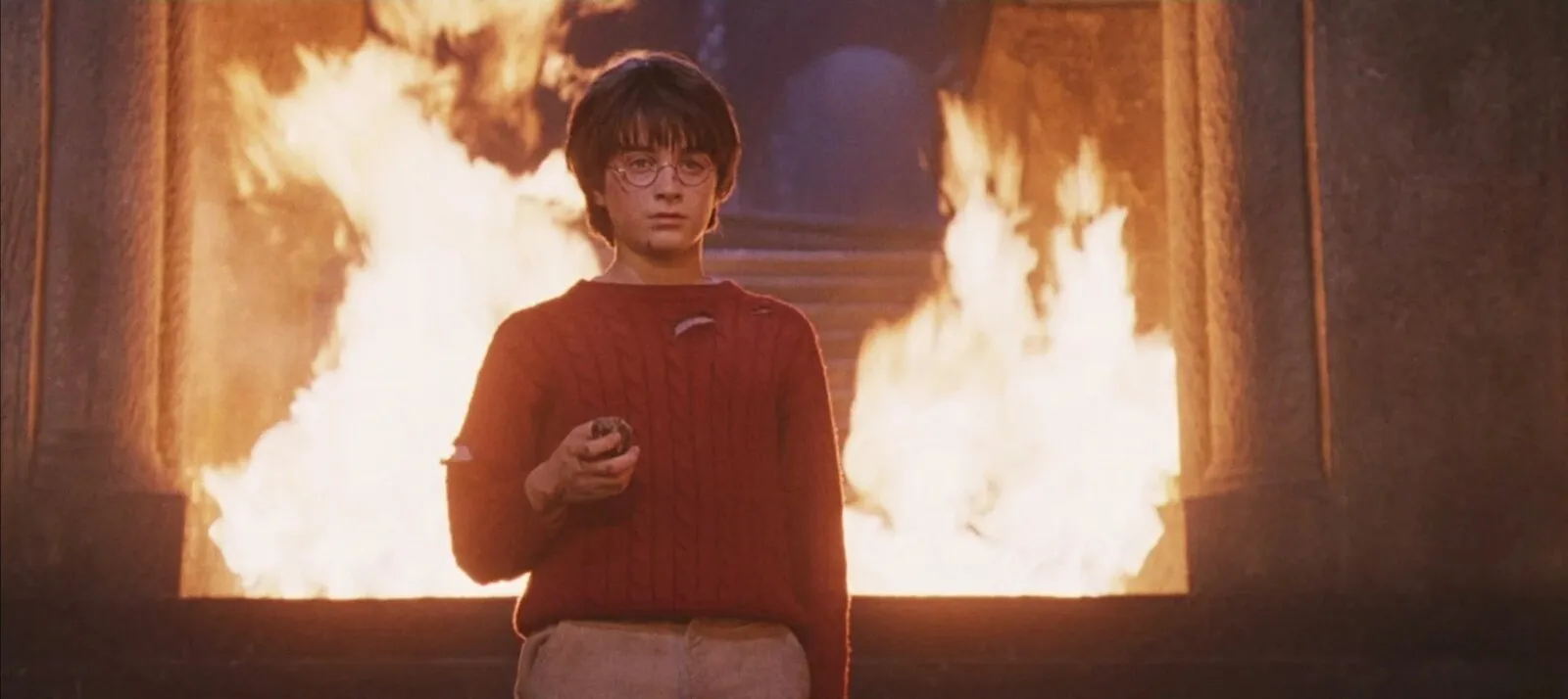 Cena do filme: Harry, de óculos, rosto arranhado e suéter vermelho rasgado, segura a pedra na mão, de pé diante de uma parede de chamas