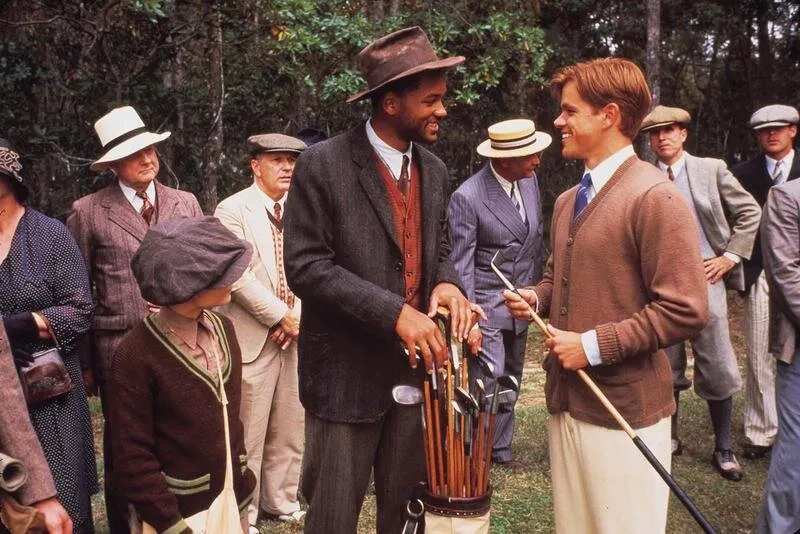Cena do filme Lendas da Vida: Will Smith, de chapéu e terno escuro, segura uma bolsa de tacos de golfe e sorri para Matt Damon, de cardigã marrom e taco na mão, cercados por homens de terno e chapéu e um menino de boina, num gramado com árvores ao fundo