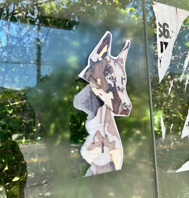 Lambe-lambe recortado de um cachorro de orelhas em pé, pintado em tons de marrom e bege, colado num vidro com árvores refletidas ao fundo
