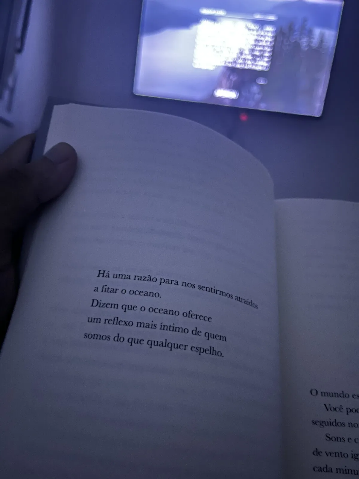 Página de um livro aberto, segurada à mão numa luz azulada, com o trecho: “Há uma razão para nos sentirmos atraídos a fitar o oceano. Dizem que o oceano oferece um reflexo mais íntimo de quem somos do que qualquer espelho.”