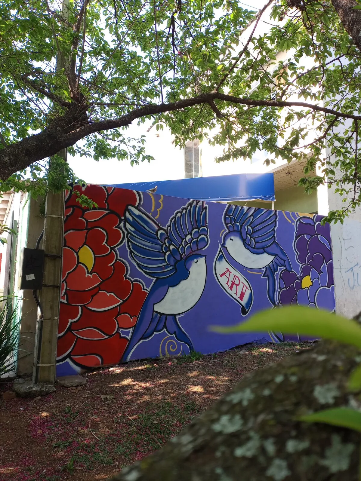 Grafite terminado num muro roxo sob uma ĂĄrvore: duas andorinhas azuis segurando uma faixa com a palavra âARTâ, entre uma flor vermelha e uma flor roxa