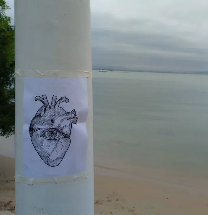 Desenho de um coração anatômico com um olho no centro, impresso numa folha colada com fita crepe num poste branco, diante de uma praia de mar calmo e céu nublado