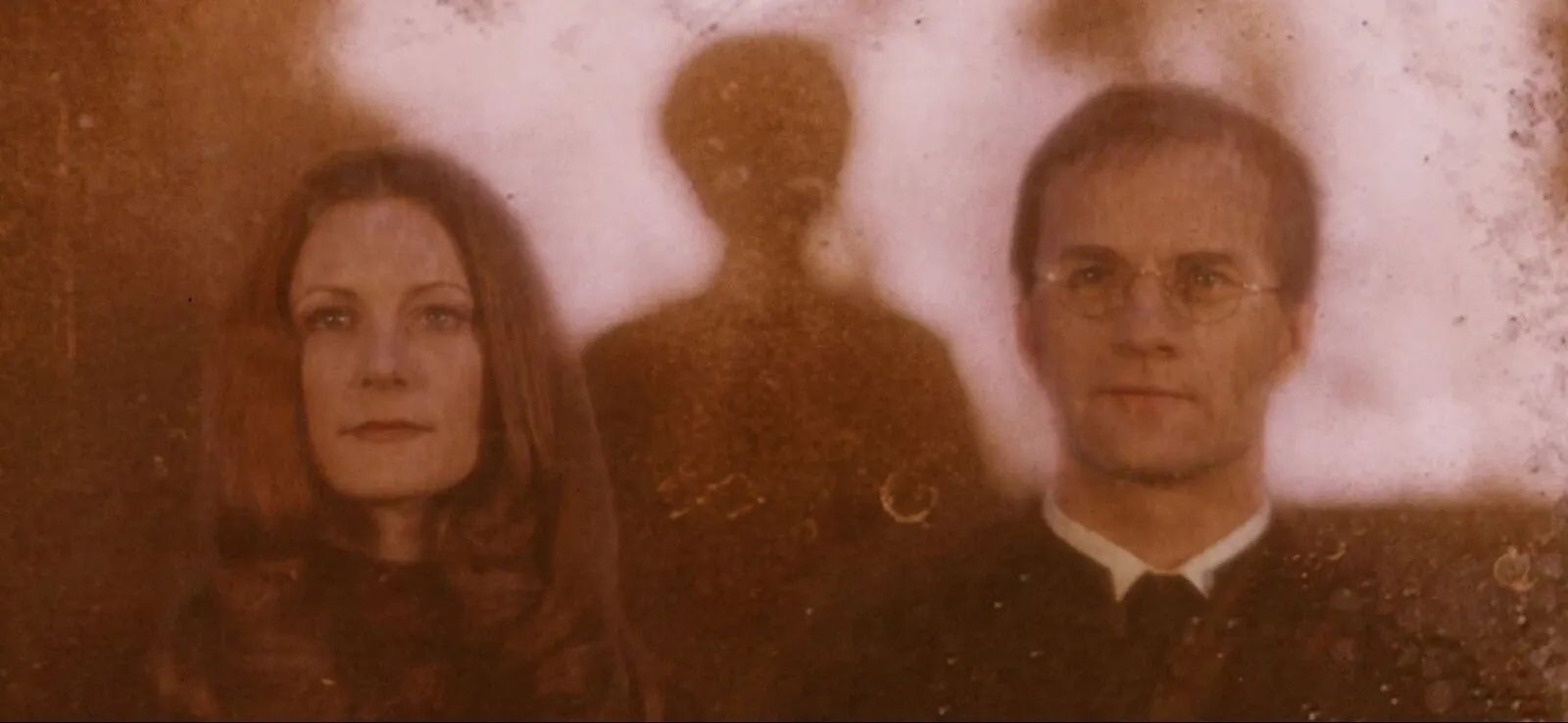 Reflexo no espelho, em tons de sépia: uma mulher ruiva e um homem de óculos olham para a frente, com a sombra de Harry entre os dois