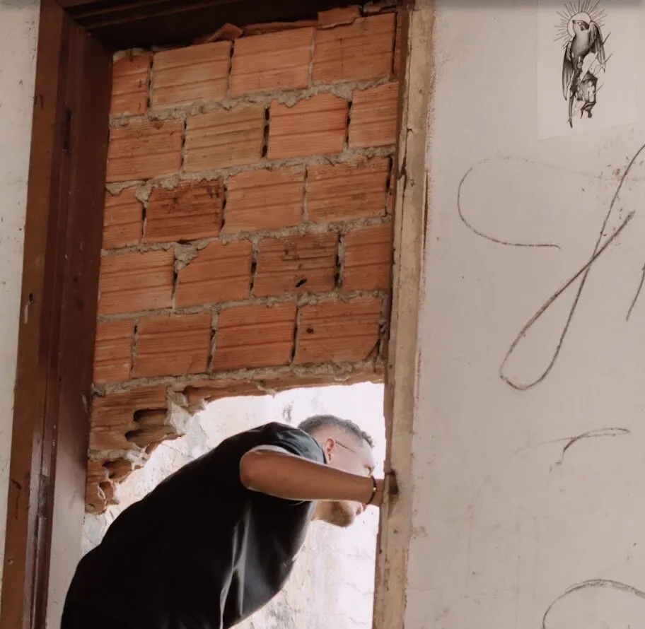 Homem de camiseta preta se curvando para atravessar um vão aberto numa parede de tijolos, numa casa em ruínas, com o desenho da andorinha colado na parede ao lado e rabiscos riscados no reboco