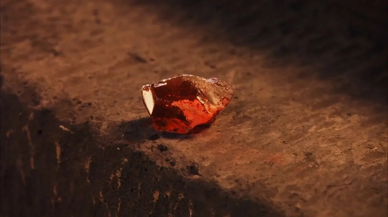 Close da Pedra Filosofal, uma pedra vermelha translúcida e irregular, sobre um degrau de pedra iluminado por uma luz alaranjada