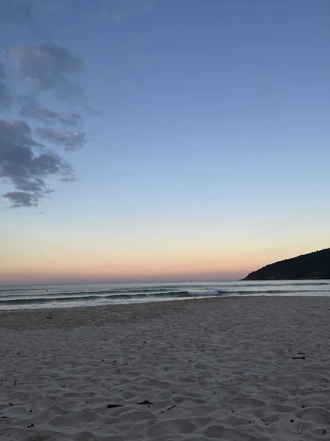 Praia ao entardecer, com o mar calmo, um morro escuro à direita e o céu azul passando para o laranja perto do horizonte