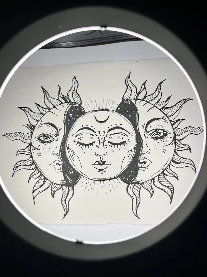 Desenho a nanquim de um sol e uma lua que se encontram: duas metades de rosto, uma de cada lado, em volta de um rosto sereno de olhos fechados, com uma lua crescente na testa