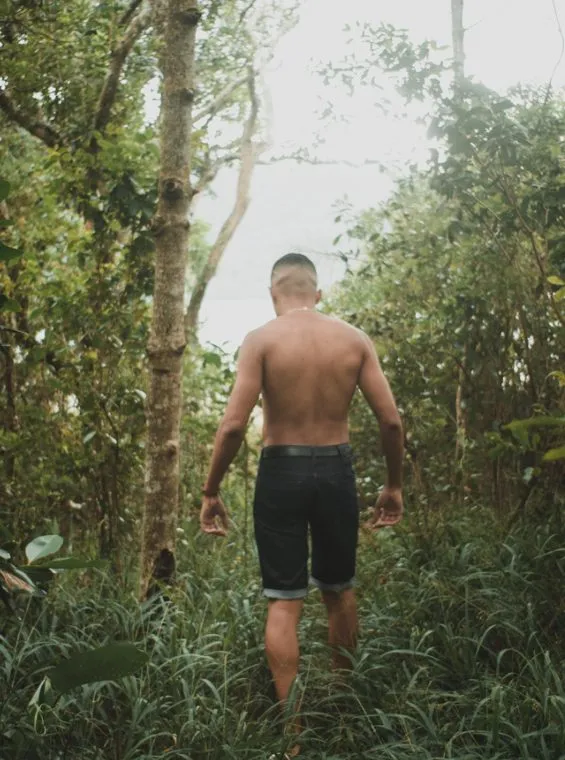 Homem sem camisa, de costas, caminhando por uma trilha estreita no meio do mato