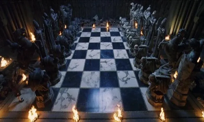 Cena do filme: um tabuleiro de xadrez gigante de casas pretas e brancas, ladeado por peças de pedra escura enormes, iluminado por pequenas chamas no chão