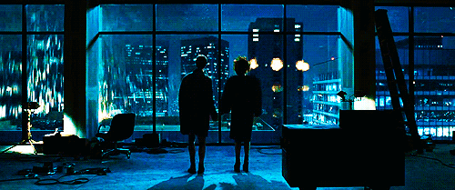 Cena final do filme: um homem e uma mulher de mãos dadas, de costas, diante de uma janela de vidro com a cidade à noite
